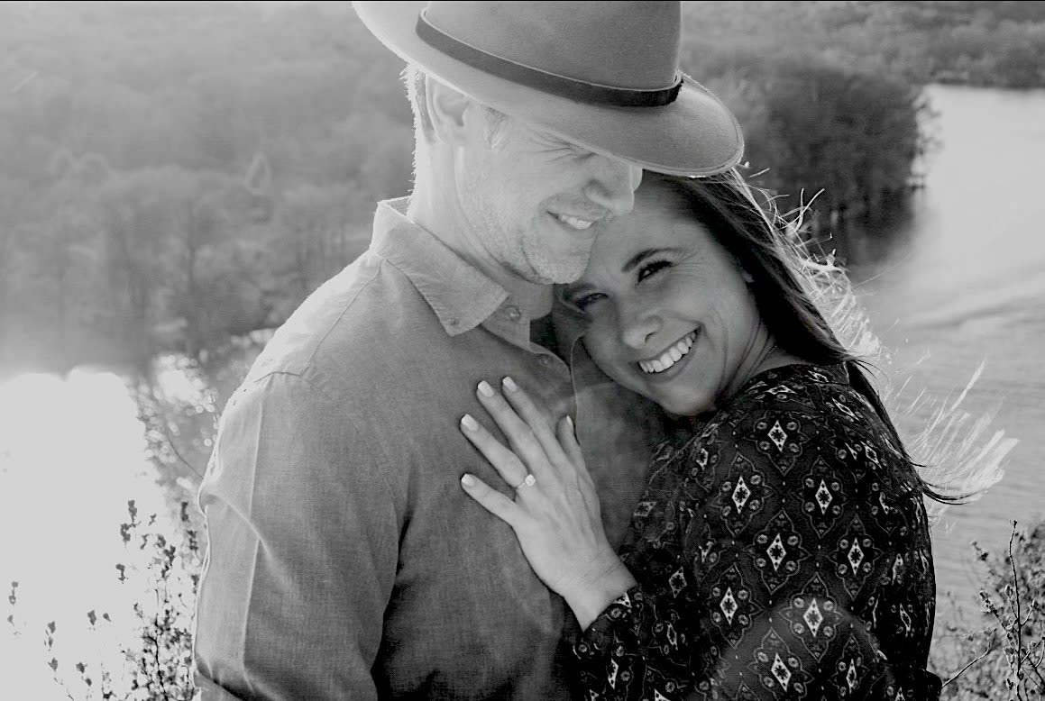 A black-and-white photo of Carly resting her head against Brandon outdoors near the water.
